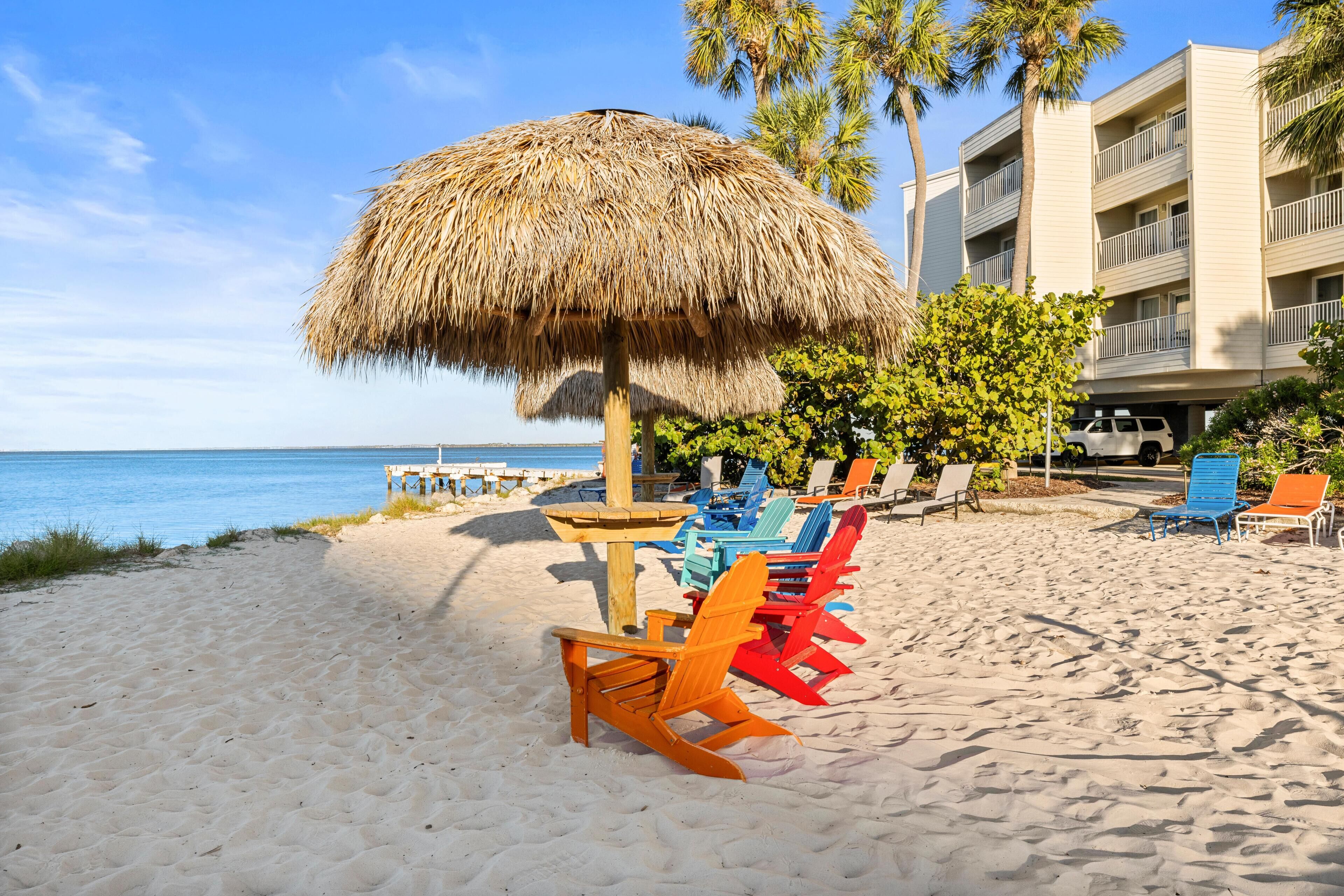 A beach with colorful Adirondack chairs, a thatched umbrella, and a building with balconies.