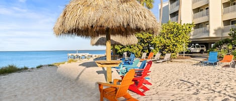 A beach with colorful Adirondack chairs, a thatched umbrella, and a building with balconies.