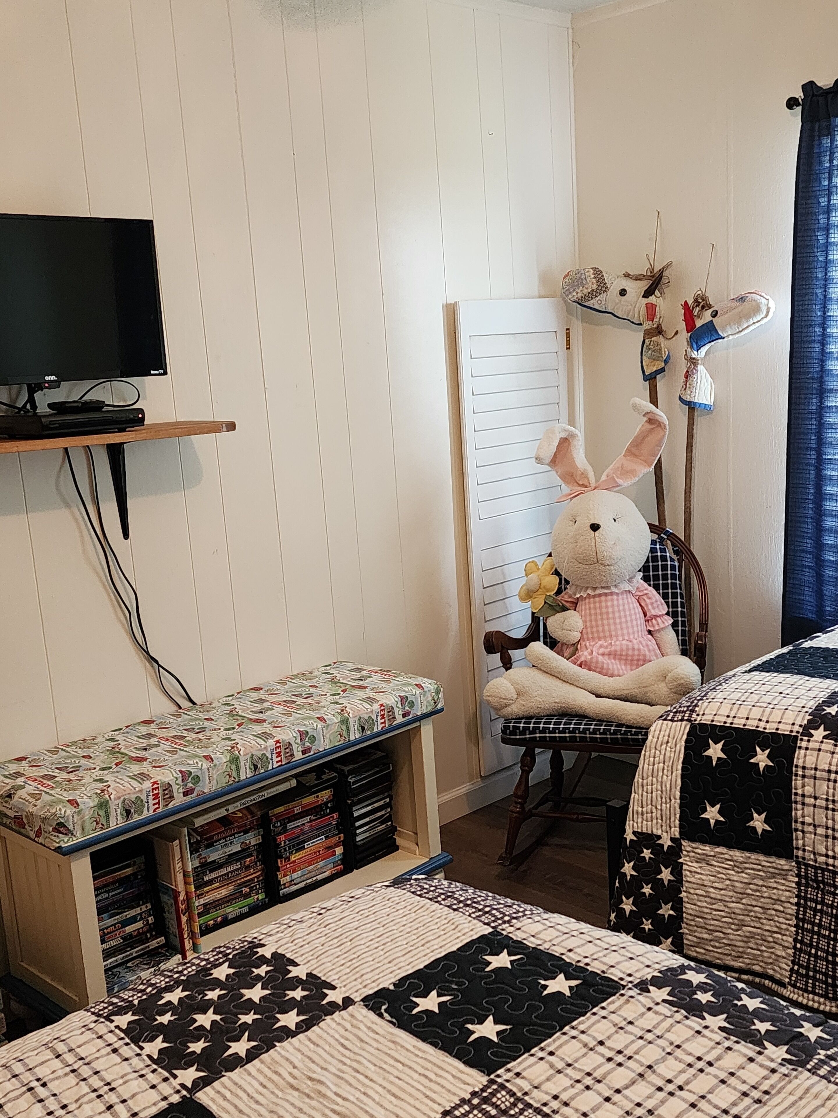 A cozy room with a television on a shelf, a plush bunny chair, a bookshelf, and a bed with a star-patterned quilt.