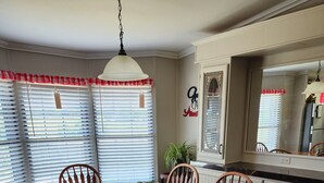 A dining room with a wooden table and chairs, a hanging light fixture, and a large window with blinds. - Welcome to our charming farmhouse retreat nestled in the Kentucky countryside. (Corinth)
