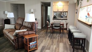 A spacious living room with a brown leather sofa, a wooden coffee table, and a dining area with a wooden table and chairs. A ceiling fan is visible, and the room has wooden flooring. - Welcome to our charming farmhouse retreat nestled in the Kentucky countryside. (Corinth)