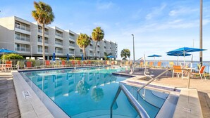 A swimming pool with lounge chairs and umbrellas, a building with balconies, and palm trees.
