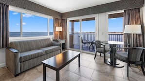A modern living room with a sofa, a coffee table, and a dining area with chairs and a table. Large windows offer a view of the sea.