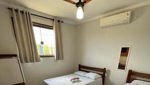 Une petite chambre propre, équipée de deux lits simples, d’une table en bois, d’un ventilateur de plafond et d’un appareil de climatisation.