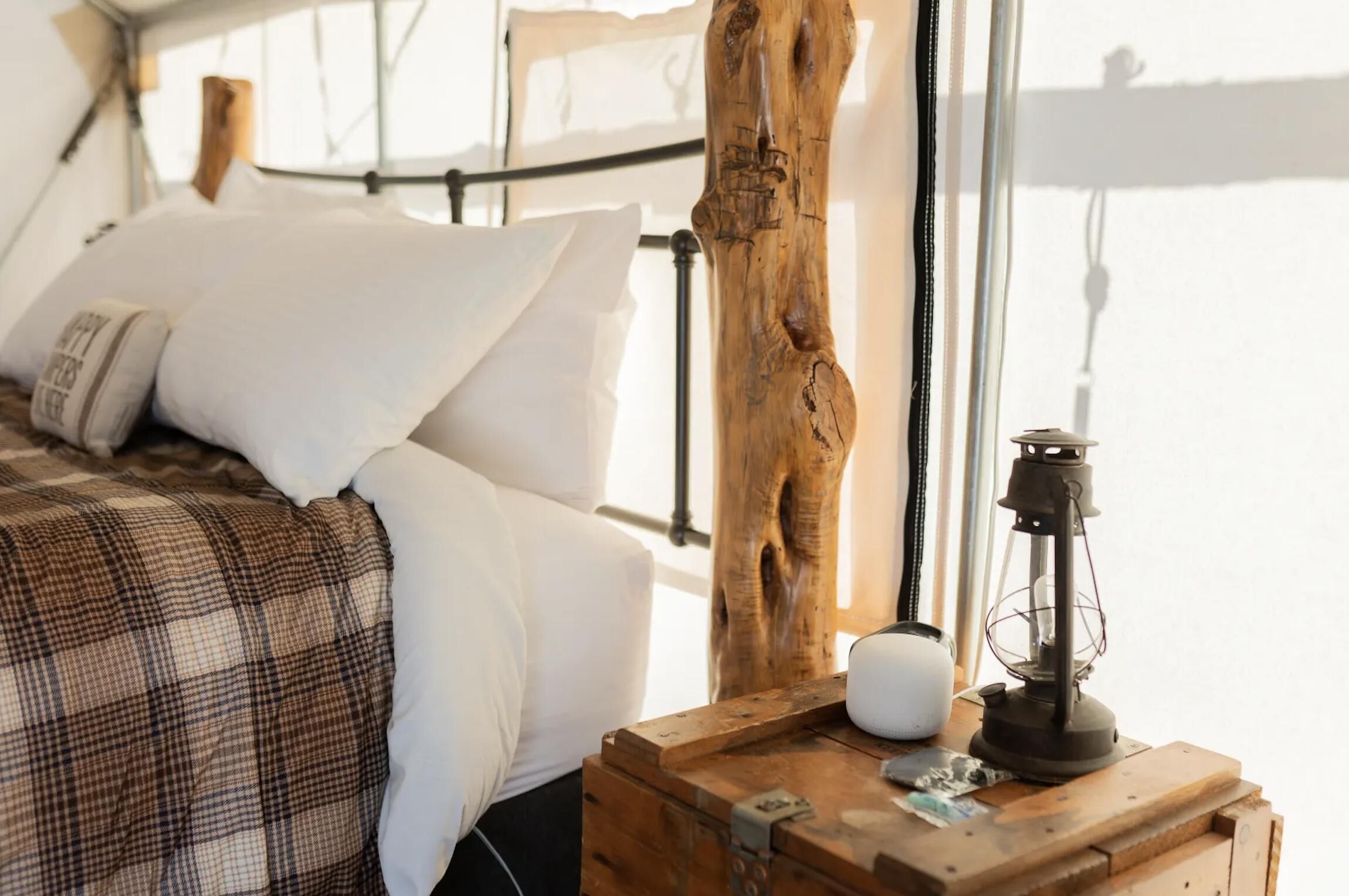 Una cama con almohadillas blancas y una manta escocesa, una mesita de noche de madera con un farol, y un tronco de árbol grande al fondo.