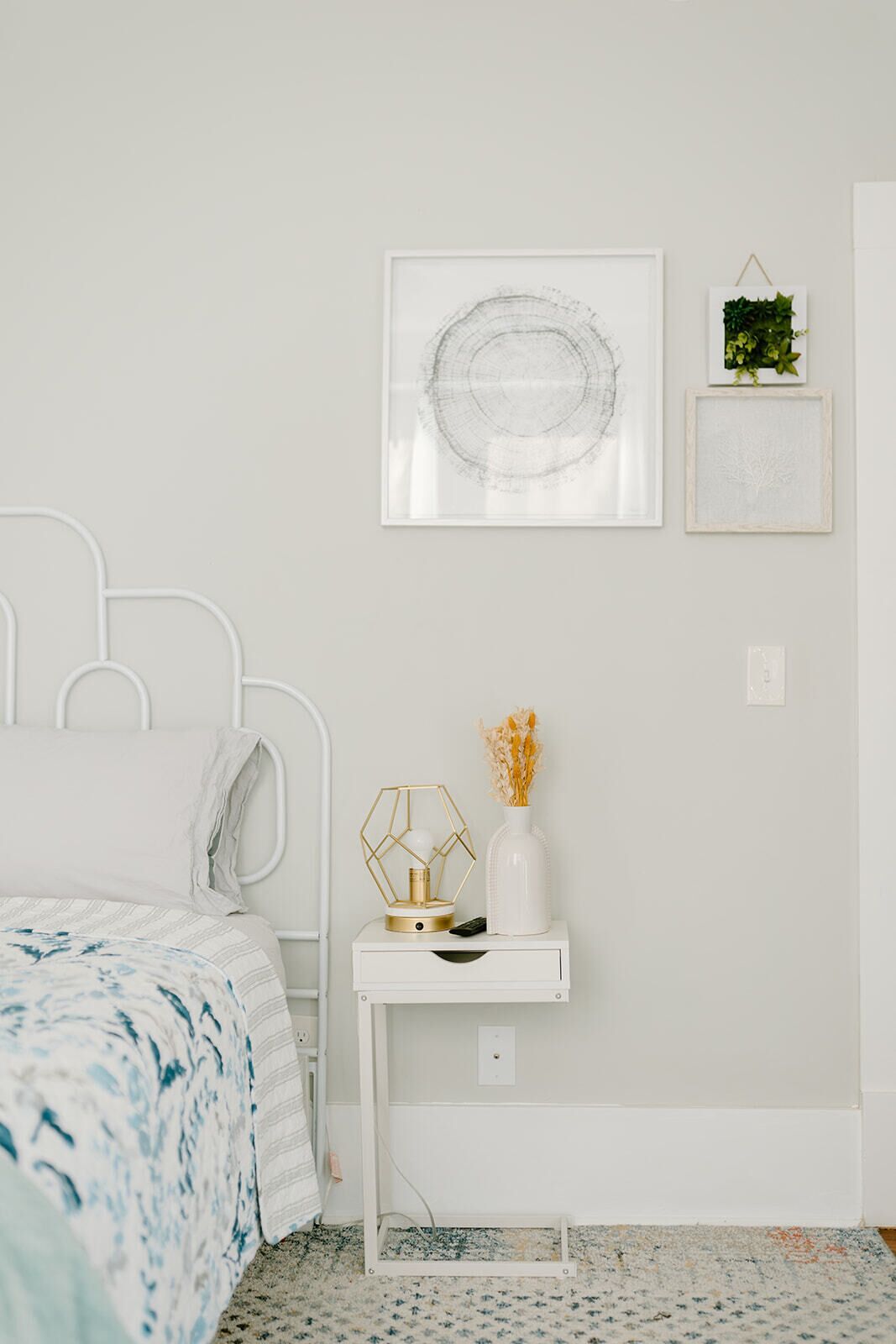 Ein Schlafzimmer mit einem weißen Bett, einem Nachttisch mit einer Vase und einer geometrischen Lampe sowie zwei gerahmten Kunstwerken an der Wand.
