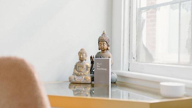 A minimalist room with a white wall, a small table with two Buddha statues, a sign reading "So, I Do Anything," and a window with a view of buildings.