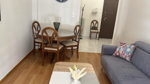 Een woonkamer met een grijze bank, een houten eettafel met vier stoelen, een vaas met gedroogde bloemen en een klok aan de muur.