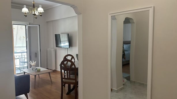 Een eetruimte met een tafel, stoelen en een kroonluchter. Er is een televisie aan de muur en een deur naar een andere kamer.