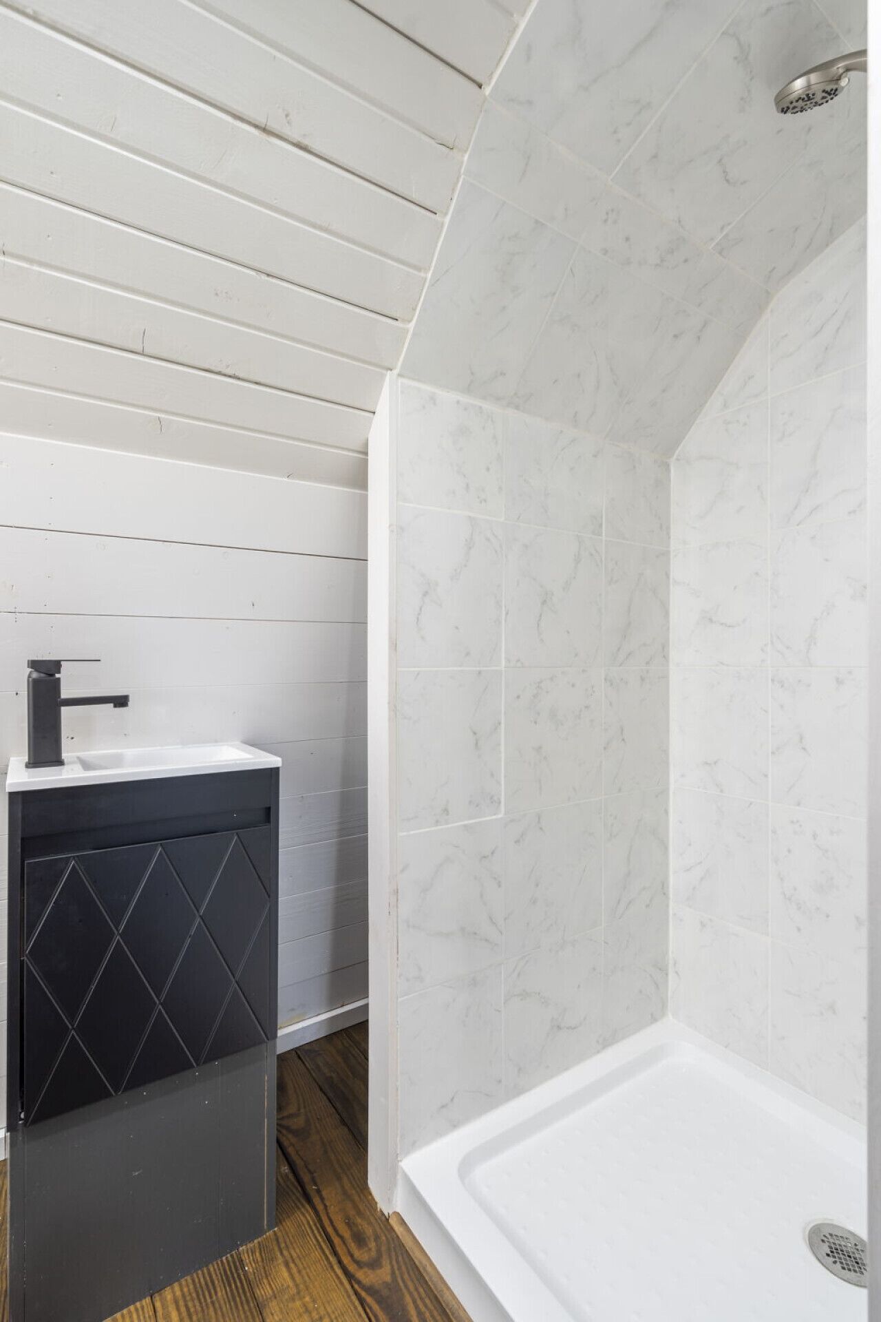 Une salle de bain avec une douche en marbre blanc, un lavabo noir et un plancher en bois.