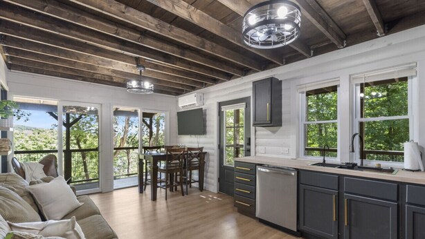 A modern kitchen with dark cabinets, a wooden ceiling, and large windows offering a view of greenery. - No Regrets at the North Fork River (Dawt)