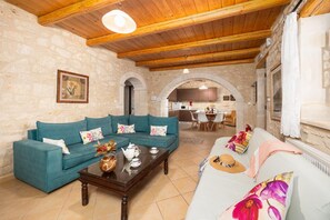 Ein gemütliches Wohnzimmer mit einem petrolfarbenen Sofa, einem Couchtisch und einem weißen Sofa mit floralen Kissen. Im Hintergrund ist eine Küche durch einen Bogen erkennbar.