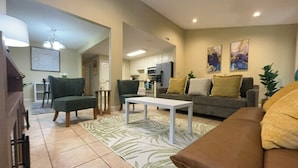 Una sala de estar con un sofá de cuero marrón, una mesa de centro blanca y un sillón verde. Se observa una cocina al fondo con armarios blancos y un refrigerador.