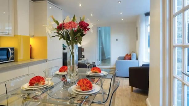 A modern kitchen with a dining table set for four, a vase with flowers, and a view to the living area. - Charming Tower Bridge House (London)