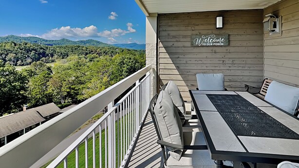 A balcony with a view of a forested landscape, a wooden wall with a 'Welcome to our porch' sign, and a table with chairs. - Beyond Blessed by Jackson Mountain Rentals (Pigeon Forge)
