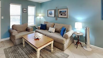 A living room with a beige sofa set, a wooden coffee table, and a patterned rug. There are two windows with blinds, a floor lamp, and a standing lamp.