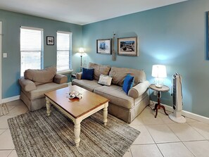 A living room with a beige sofa set, a wooden coffee table, and a patterned rug. There are two windows with blinds, a floor lamp, and a standing lamp. - Kontiki Beach #221 by Miss Kitty’s (Rockport)