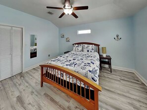 A bedroom with a bed, a ceiling fan, a nightstand, a lamp, and a window. - Our Shore Thing by Ryson Vacation Rentals (Galveston)