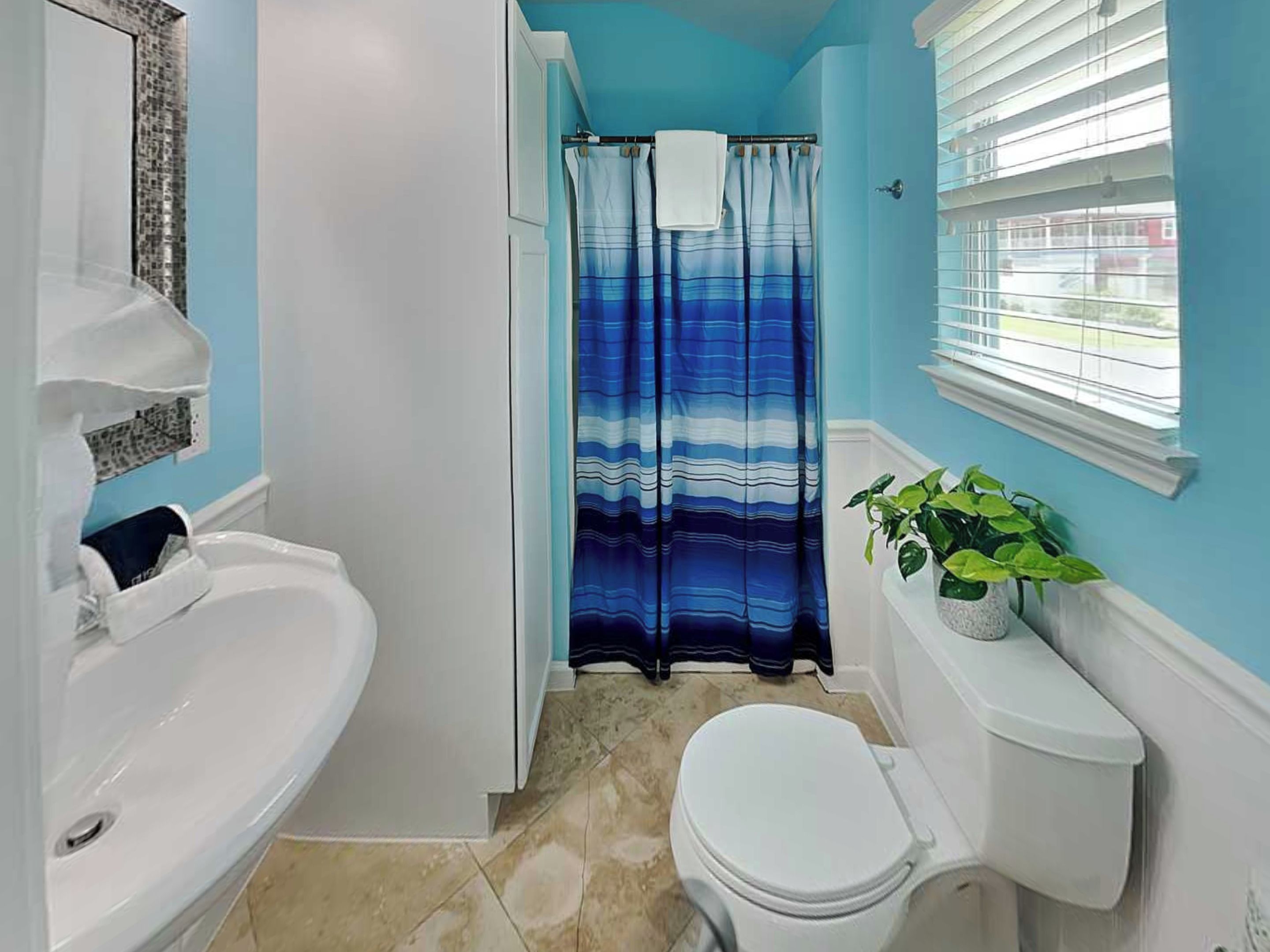 A bathroom with a white sink, a toilet, a shower curtain, and a window with blinds.