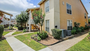 A two-story building with a beige exterior, a covered outdoor area with stairs, and a well-maintained garden with various plants and flowers. - Glowing Fun by Ryson Vacation Rentals (Galveston)