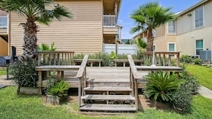 A wooden deck with steps leading to a balcony, surrounded by palm trees and a well-maintained garden. - Glowing Fun by Ryson Vacation Rentals (Galveston)