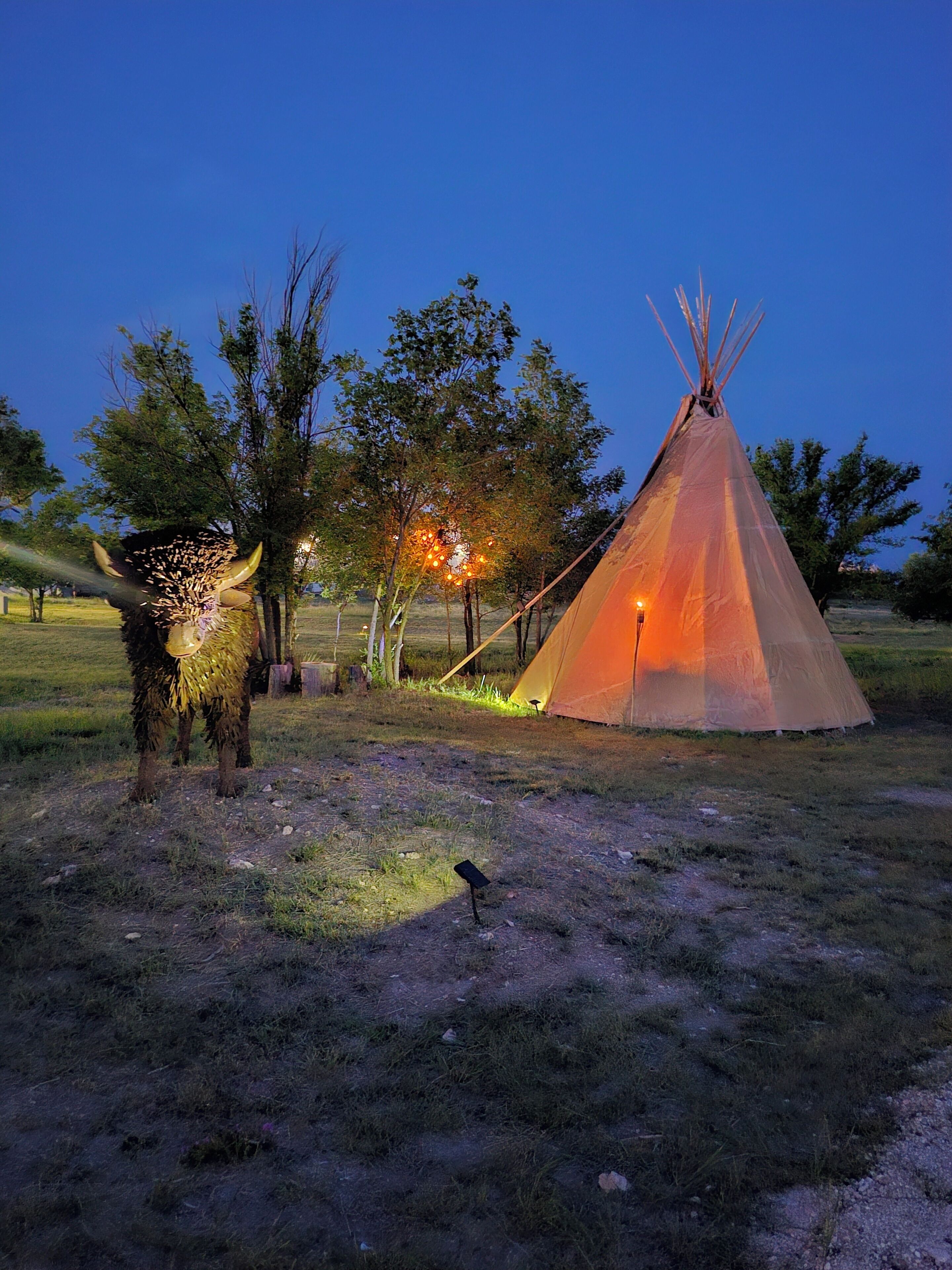 A tipi with a light source inside, surrounded by trees and grass.