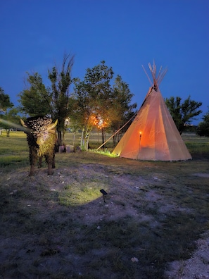 Un tipi con una fuente de luz en su interior, rodeado de árboles y hierba.