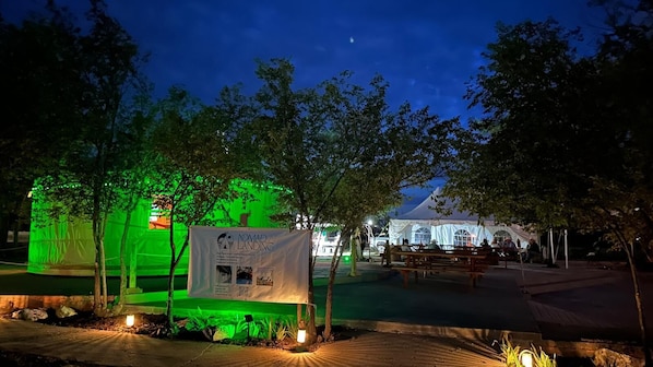 Escena nocturna al aire libre con una tienda iluminada en verde y un cartel que dice 'Nomad Landing'.
