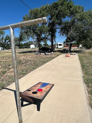 Un camino de concreto con una mesa de madera y estructura de metal, rodeado de árboles y vehículos recreativos.