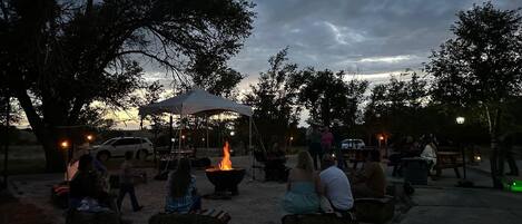 Reunión al aire libre con gente sentada alrededor de un brasero bajo una pérgola, con árboles y un atardecer al fondo.