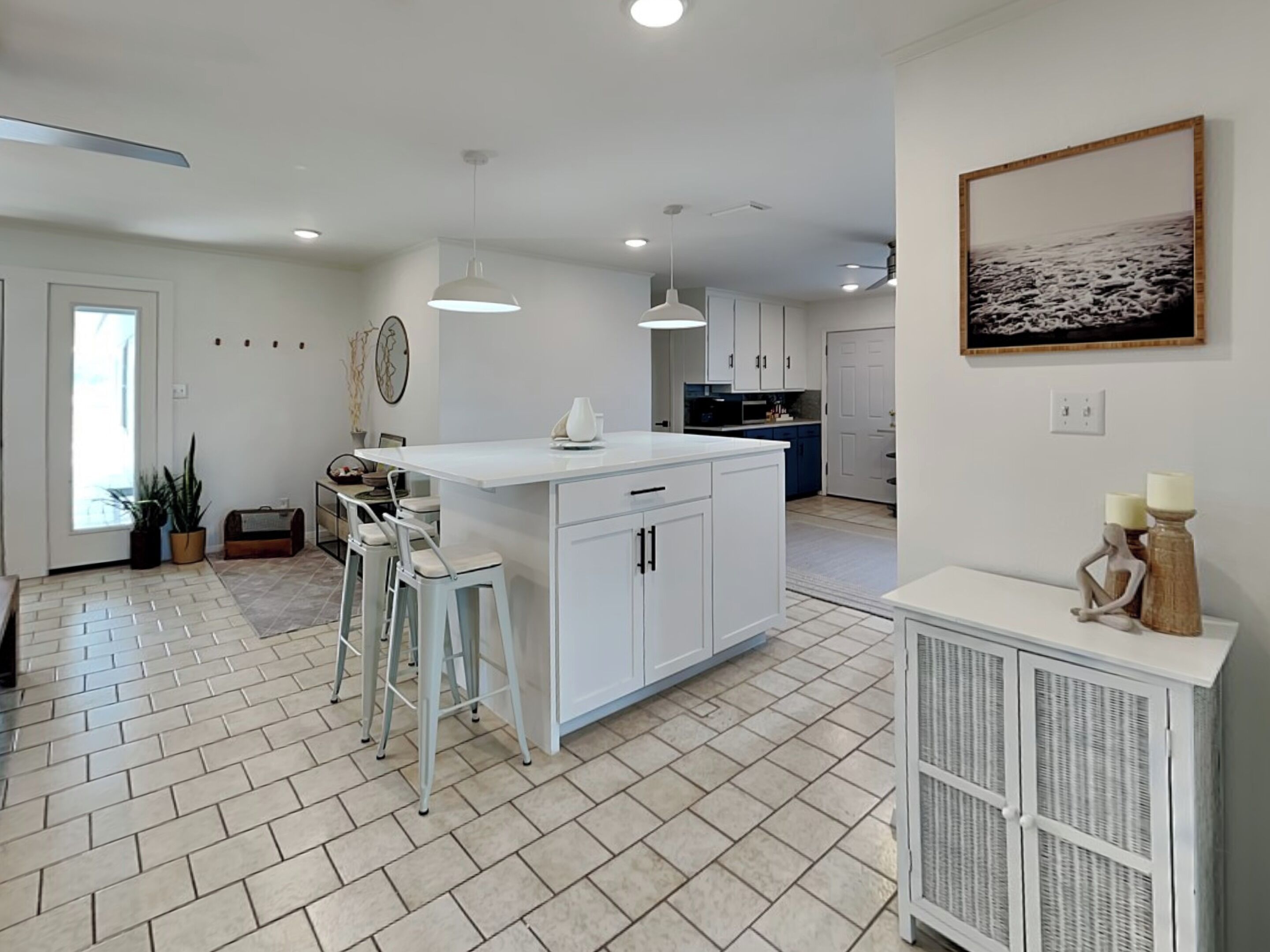 A modern kitchen with a central island, bar stools, pendant lighting, and a dining area with a view of the sea.