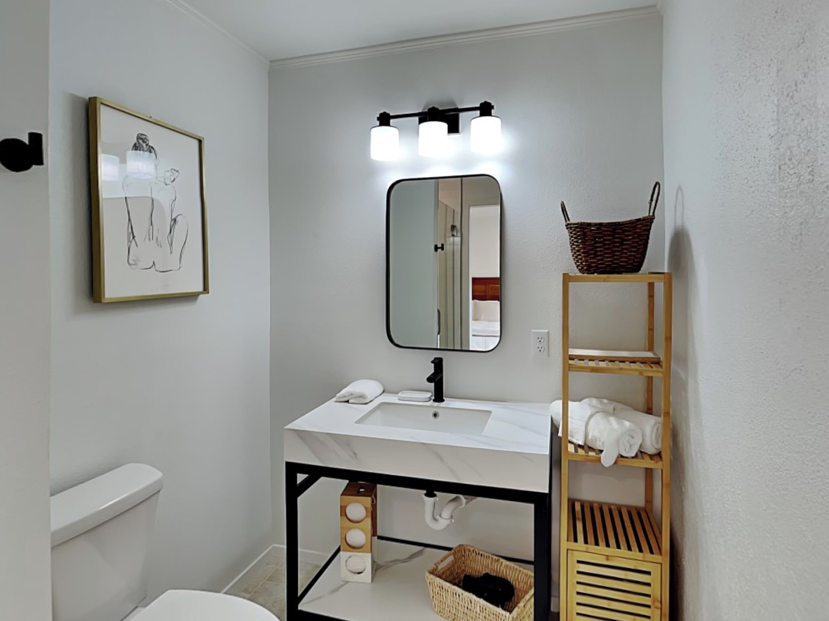 A modern bathroom with a white sink, a mirror, and a toilet. There is a framed artwork on the wall and a shelf with towels.