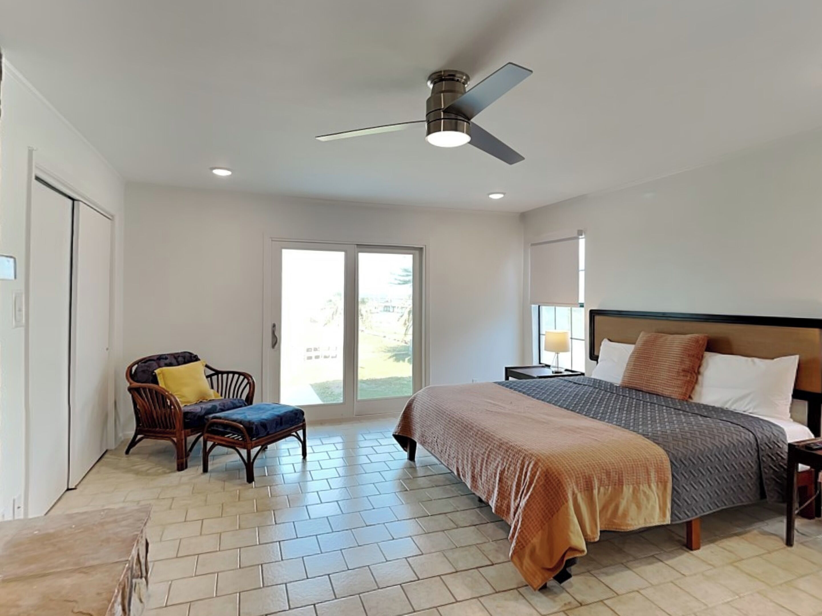 A bedroom with a bed, a chair, a table, a ceiling fan, and a view through a glass door.