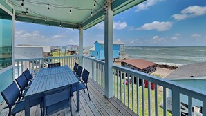 Een overdekt terras buiten met een tafel en stoelen, met uitzicht op een strand met huizen en de oceaan.