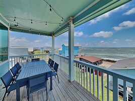 Una terraza techada al aire libre con una mesa y sillas, con vistas a una playa con casas y el océano.