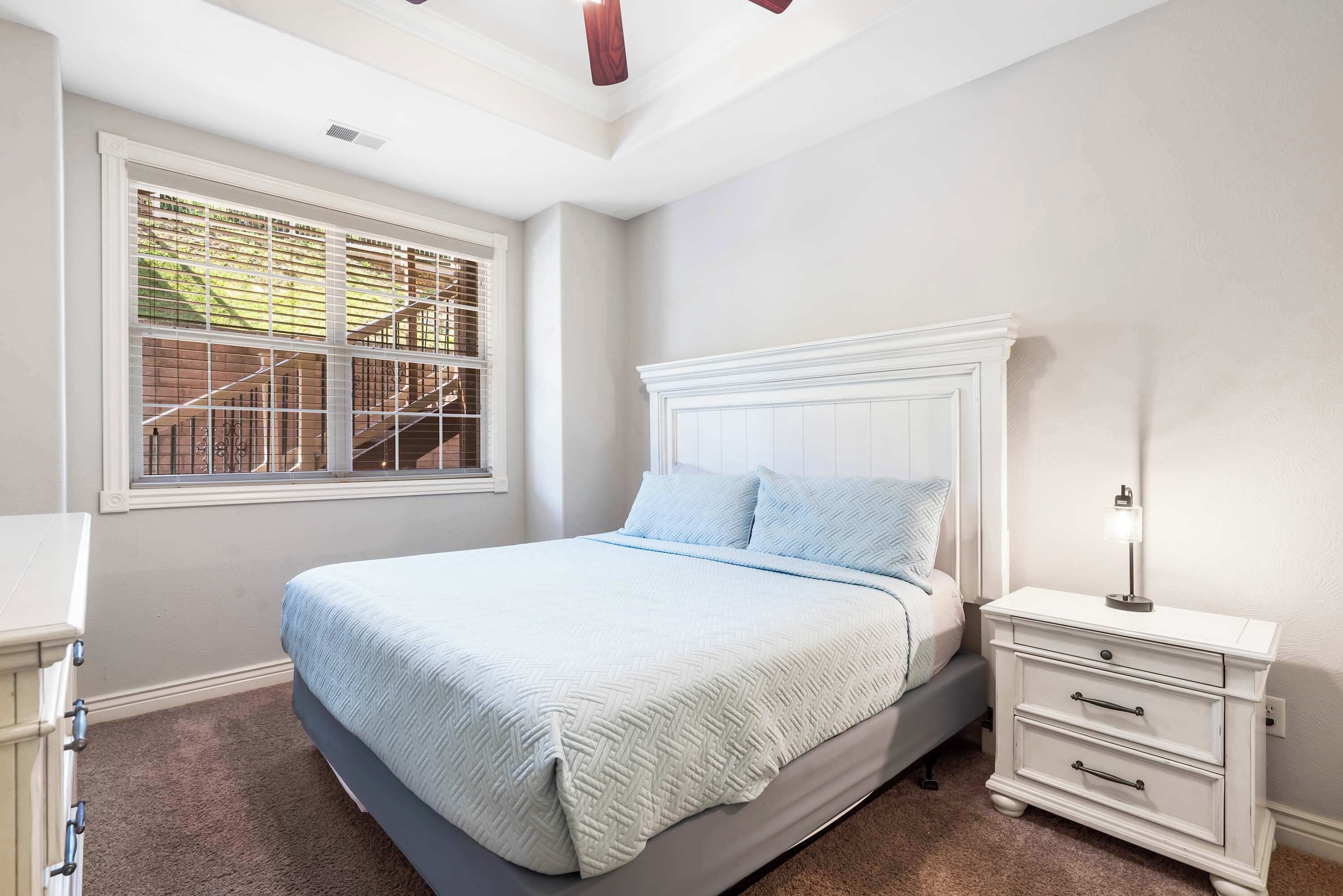 A bedroom with a bed, a nightstand, a window with blinds, and a ceiling fan.