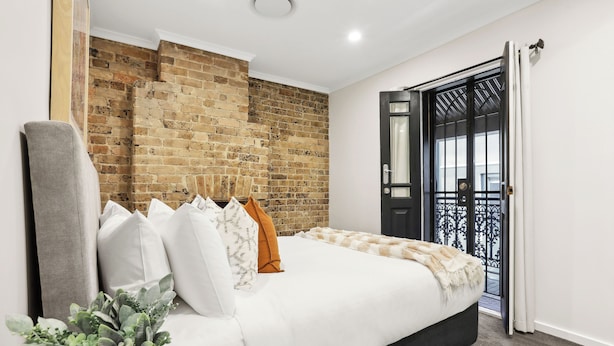 Une chambre avec un mur en briques, un lit avec une literie blanche et orange, un fauteuil gris et une plante en pot.