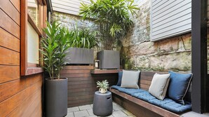 Un espace extérieur accueillant, avec un banc en bois et des coussins, entouré de plantes en pot et d’un mur de pierres.