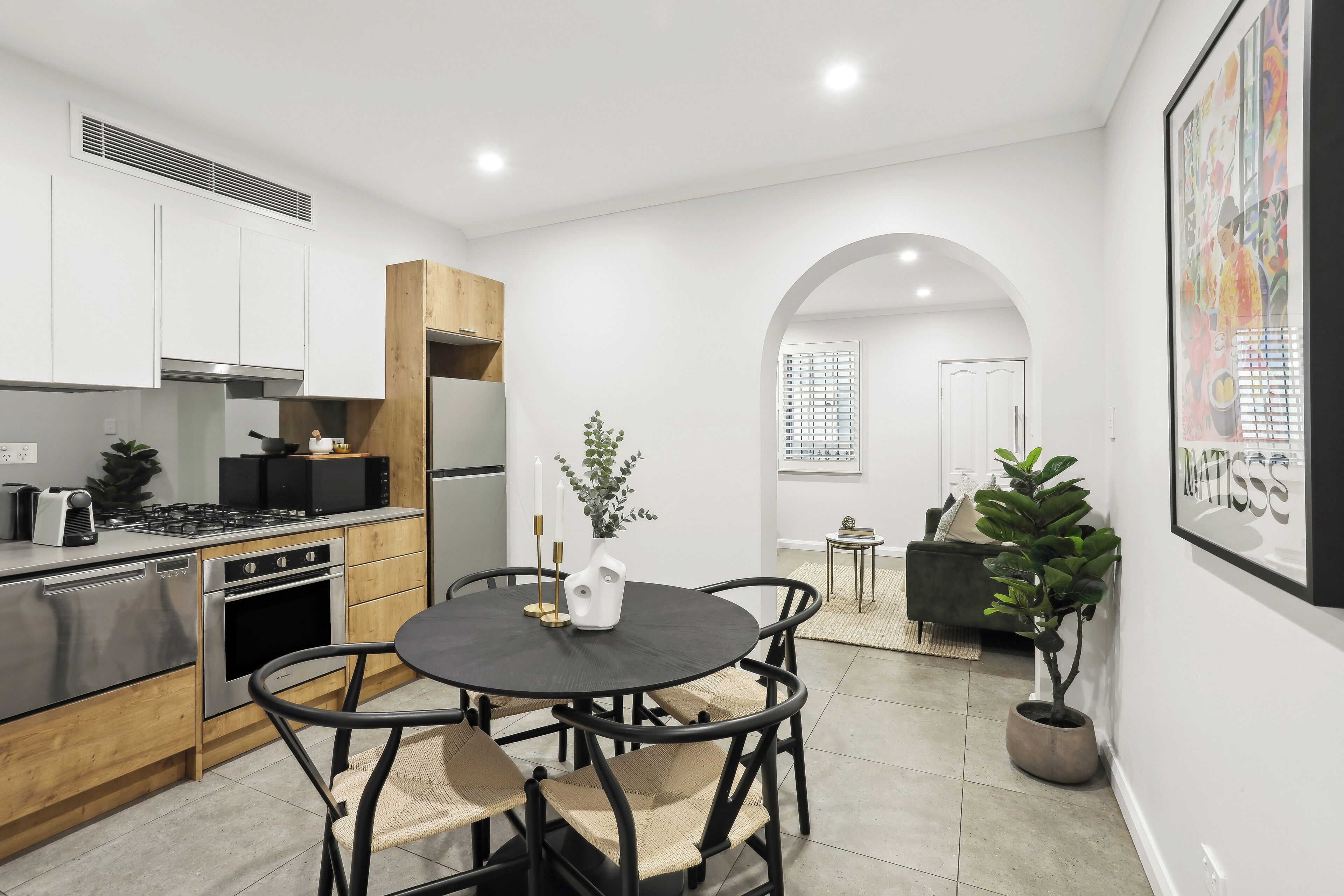 A modern kitchen with white cabinetry, a dining area with a round table and chairs, and a living space with a sofa and plants.