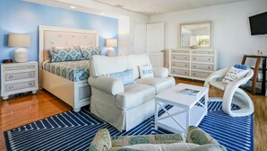 A bedroom with a bed, a white sofa, a white armchair, a small white table, a blue and white striped rug, a ceiling fan, and a dresser with drawers. - Sunshine Lane by Distinctive Beach Rentals (Treasure Island)