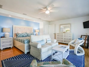 A bedroom with a bed, a white sofa, a white armchair, a small white table, a blue and white striped rug, a ceiling fan, and a dresser with drawers.