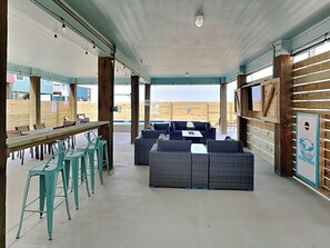 A covered outdoor seating area with a long table and chairs, a sofa, and a TV mounted on the wall. - The Blue Heron by Silver Sands Vacation Rentals (Port Aransas)