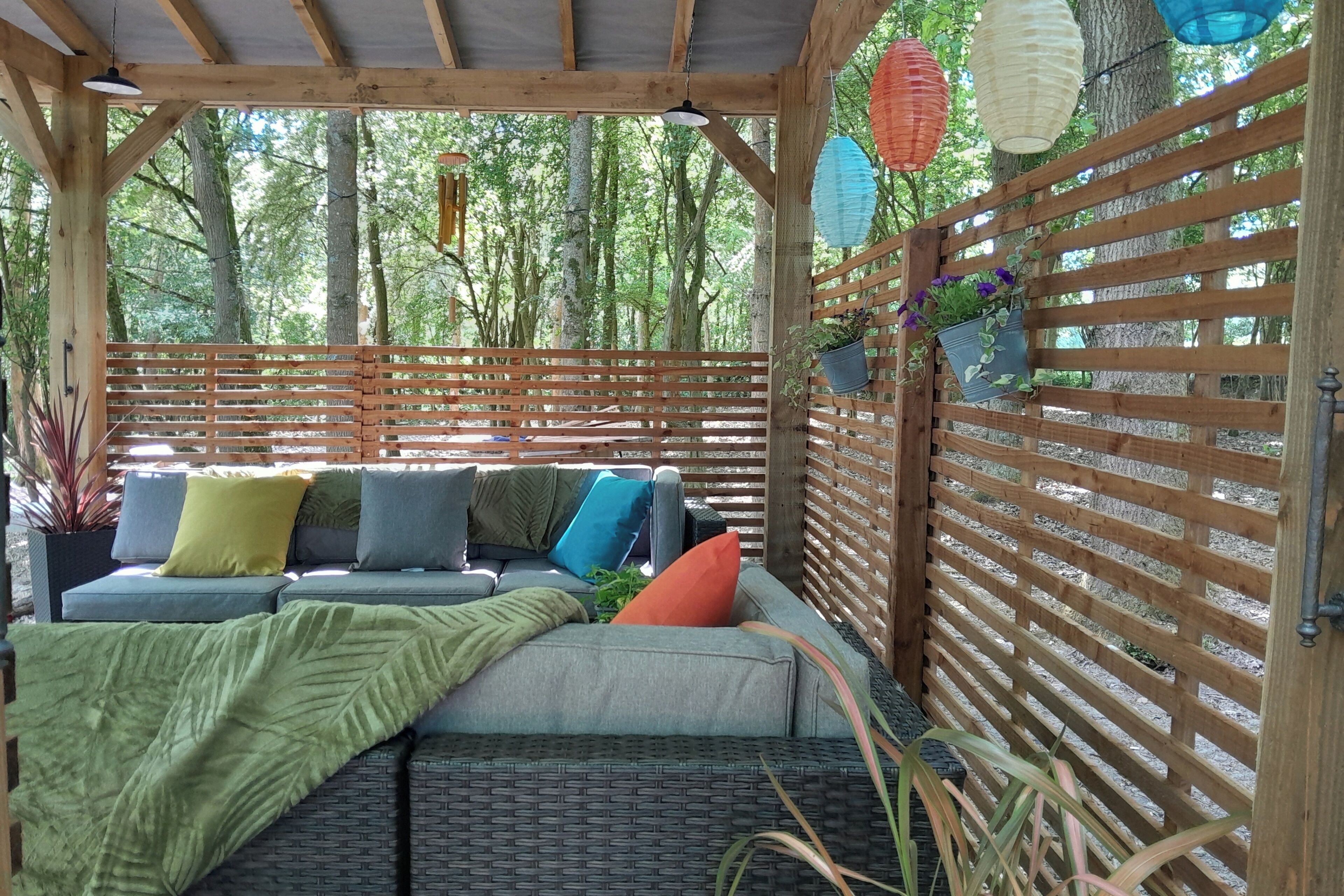 A covered outdoor seating area with a wooden bench and cushions, surrounded by a wooden fence and potted plants.