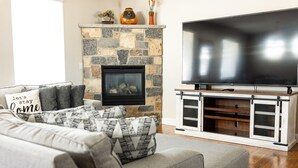 Ein Wohnzimmer mit Kamin aus Stein, einem grauen Sofa mit gemusterten Zierkissen und einem Flachbildfernseher auf einem Holzständer.
