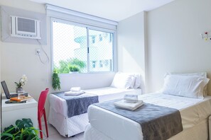 Egy kétágyas szoba, íróasztallal és laptopal felszerelve, ablakból zöld növényzetre nyíló kilátással, és légkondicionálóval.