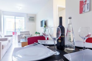 Egy modern, jól megvilágított szobában étkezőasztal van, melyen tányérok, borospoharak és palackok találhatók. A háttérben egy piros szék és egy televízió látható.