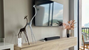 Ett modernt vardagsrum med en väggmonterad TV, ett träbord, en girafffigur, en vas med torkade växter och en balkong med en stol.