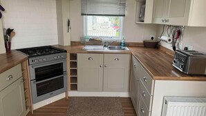 A compact kitchen with a gas stove, oven, and a toaster. There is a window with blinds, a sink, and a radiator. - Heacham Beach Caravan-sleeps 6-pets Ok-parking (King's Lynn)