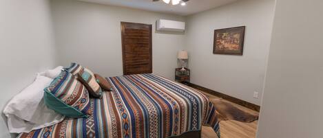 Un dormitorio con cama, ventilador de techo, equipo de aire acondicionado, mesita de noche con lámpara y un cuadro en la pared.