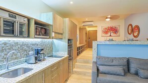 A modern kitchen with a built-in oven, microwave, and coffee maker. There is a sink with a faucet, and a countertop with a toaster and a coffee pot. A gray sofa is in the living area, and a painting of a tree is on the wall. - Studio with Roof Pool Near Beach (Playa del Carmen)